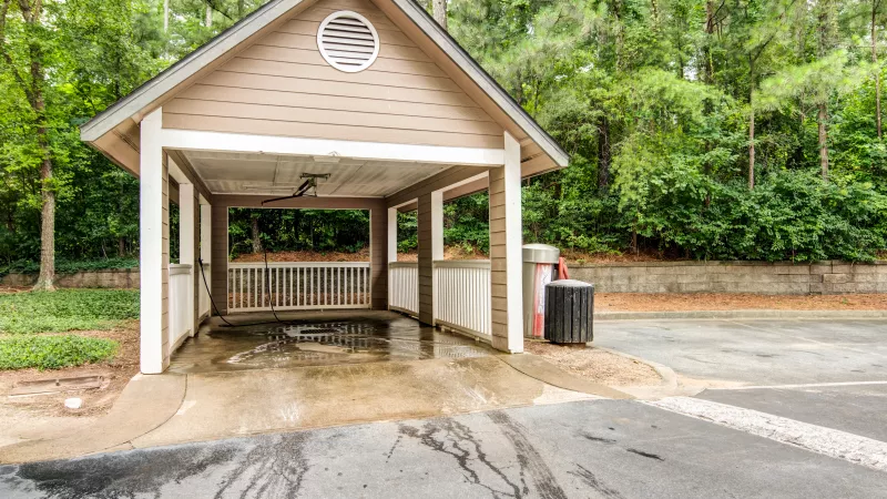 Tan wooden pavilion with a gabled roof, covering a wet concrete pad next to a road, surrounded by trees.