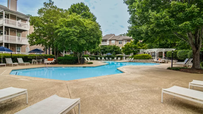 Outdoor swimming pool area with lounge chairs, apartment buildings, and lush green trees.