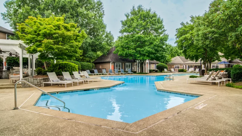 Outdoor swimming pool with light blue water and lounge chairs, surrounded by green trees and buildings.