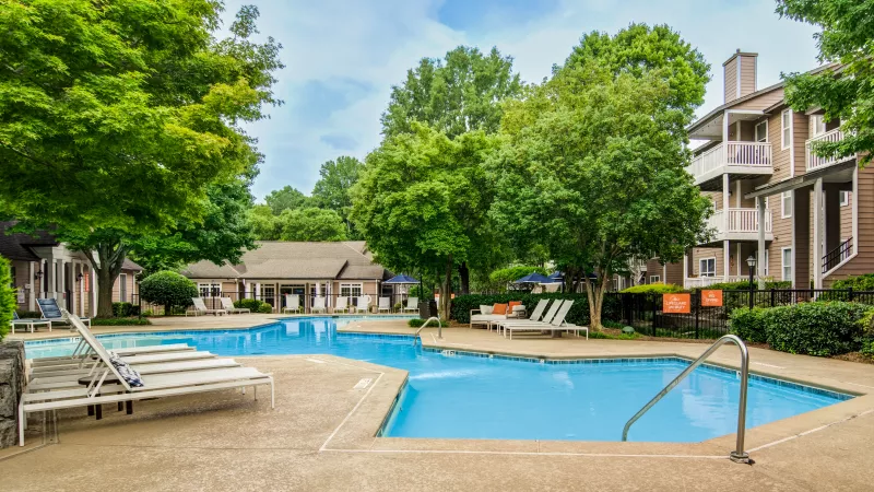 Outdoor swimming pool surrounded by lounge chairs, lush trees, and apartment buildings under a partly cloudy sky.