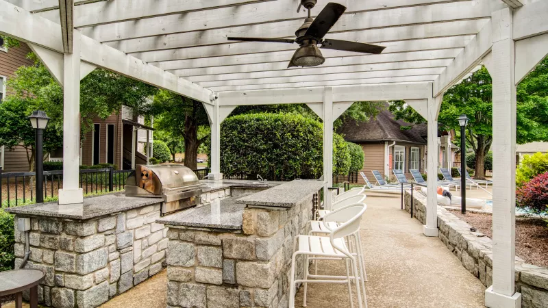 Outdoor kitchen and bar under a white pergola, with stone counters, a grill, bar stools, next to a pool area.