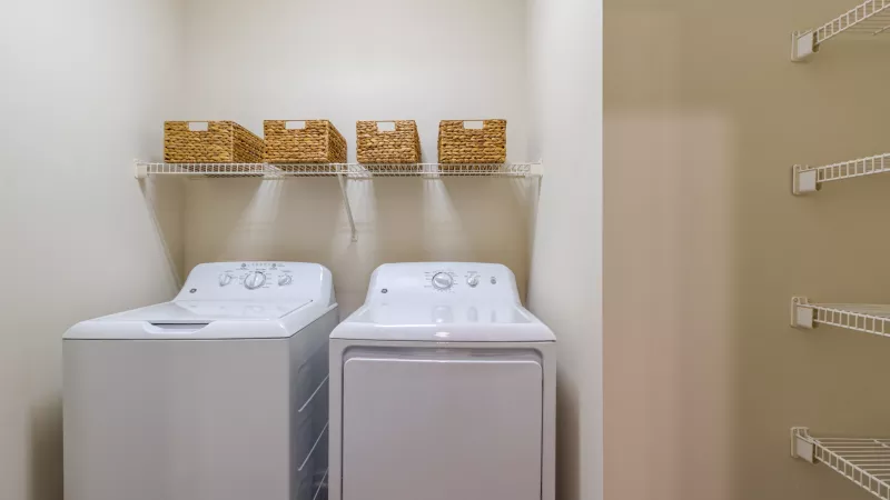 White washer and dryer in a bright laundry area with a shelf of woven baskets. Shelving unit visible on the right.