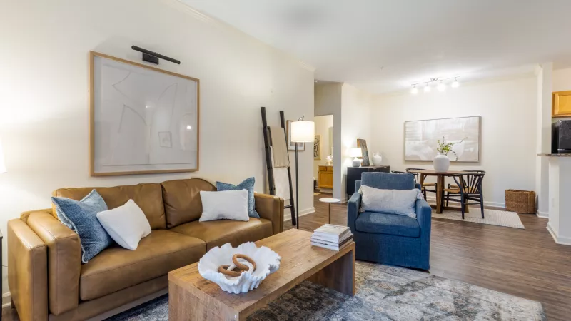 Photograph of a modern living room with a tan leather sofa, blue armchair, wooden coffee table, and an abstract rug. Dining area in background.