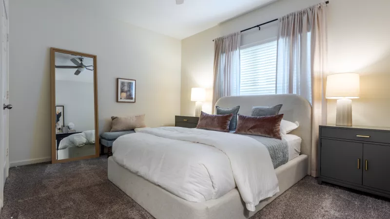 Spacious, modern bedroom with a bed, neutral bedding, blue &amp; rust pillows, dark nightstands, and a full-length mirror.