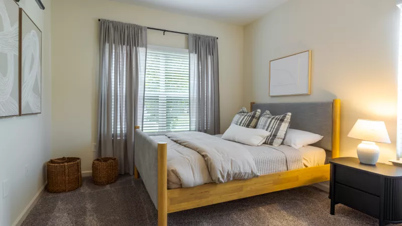 Bedroom with light wood bed, gray headboard, neutral bedding, black nightstand with lamp, and sheer gray curtains.