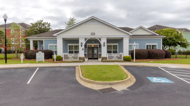 Photograph of a light blue clubhouse with white trim, a paved parking lot, grass, and shrubs under an overcast sky.