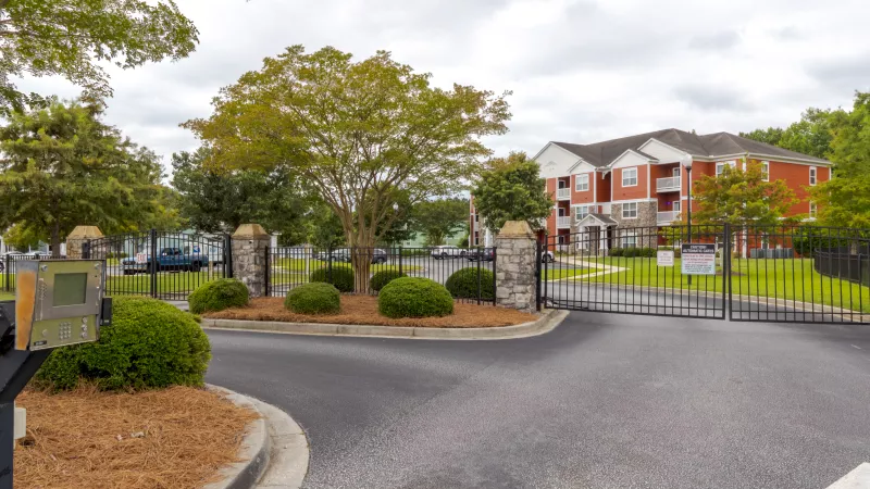 Gated entrance to an apartment complex with open black metal gates, stone pillars, and an orange apartment building in the background.