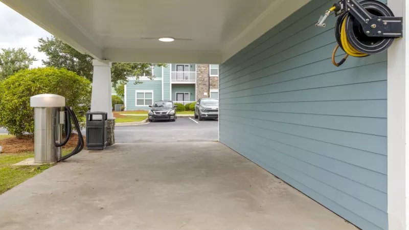 Covered car vacuum station and yellow hose reel next to a blue-sided building. Apartments and cars are in the background.