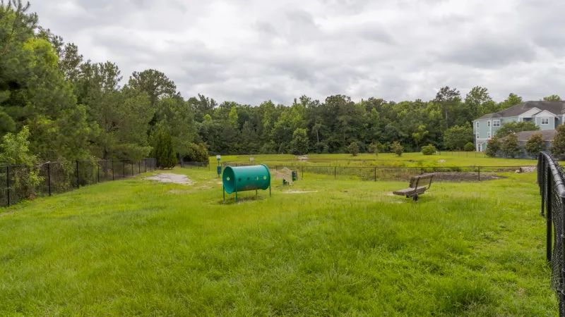 A fenced outdoor dog park with green grass, play equipment, and a wooden bench. Trees and a building in the background.