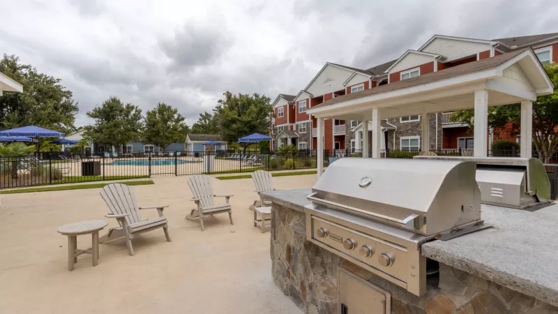 Outdoor grilling station with a large stainless steel grill on a stone counter, patio, pool, and apartment building under a cloudy sky.