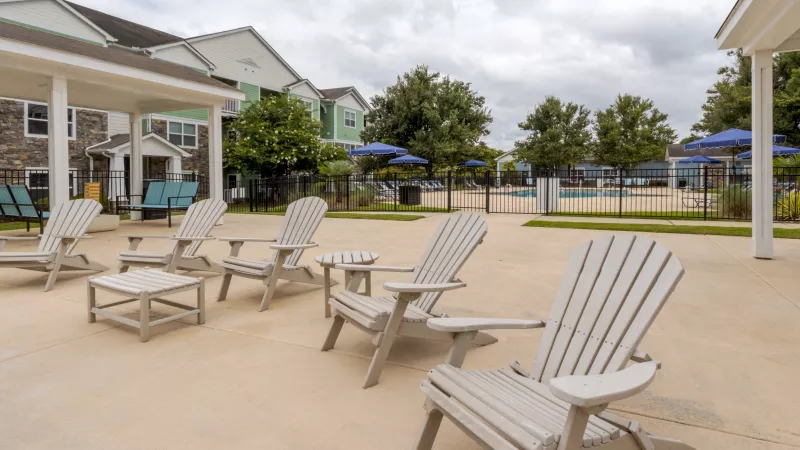 Outdoor patio with light Adirondack chairs and tables. Apartment buildings and a pool with blue umbrellas in the background.