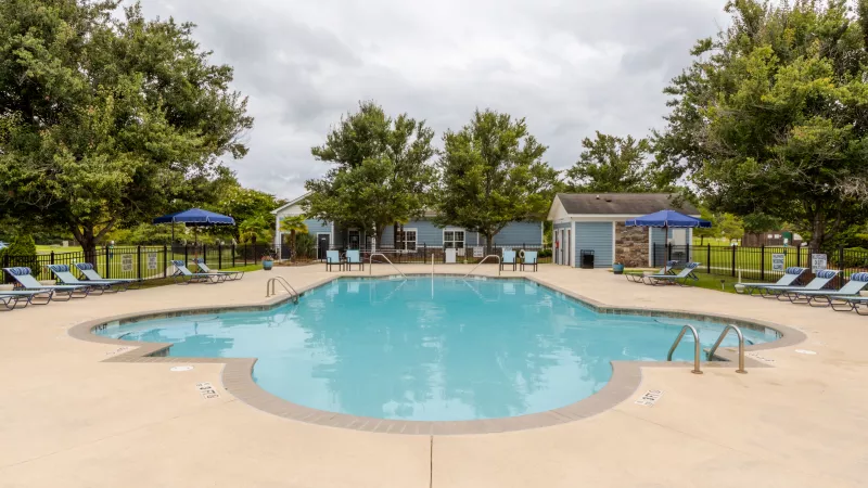 Outdoor swimming pool with blue lounge chairs and umbrellas, surrounded by trees and light blue buildings under a cloudy sky.