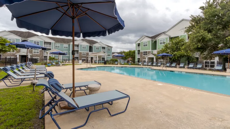Residential swimming pool with blue lounge chairs, umbrellas, and apartment buildings on a cloudy day.