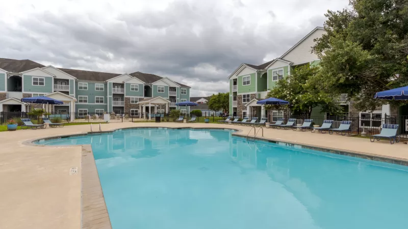Outdoor swimming pool with lounge chairs, flanked by light blue and green apartment buildings under an overcast sky.