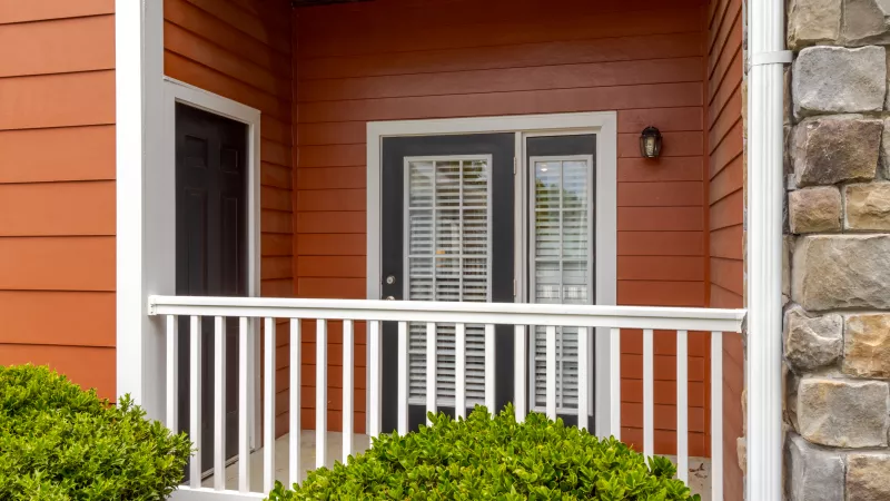 Red-orange building exterior with white balcony railing, green bushes, and two dark doors, one with glass panes.