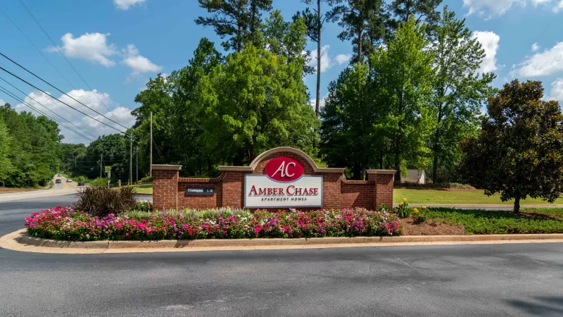 Amber Creek Apartment Homes sign: brick arch with a red AC logo, surrounded by pink flowers under a blue sky.