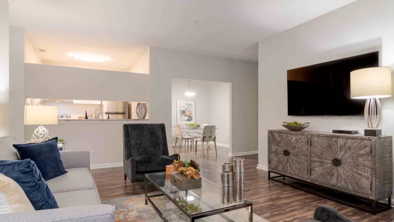 Modern living room with gray couch, dark armchairs, glass coffee table, ornate TV stand, and dining area in background.