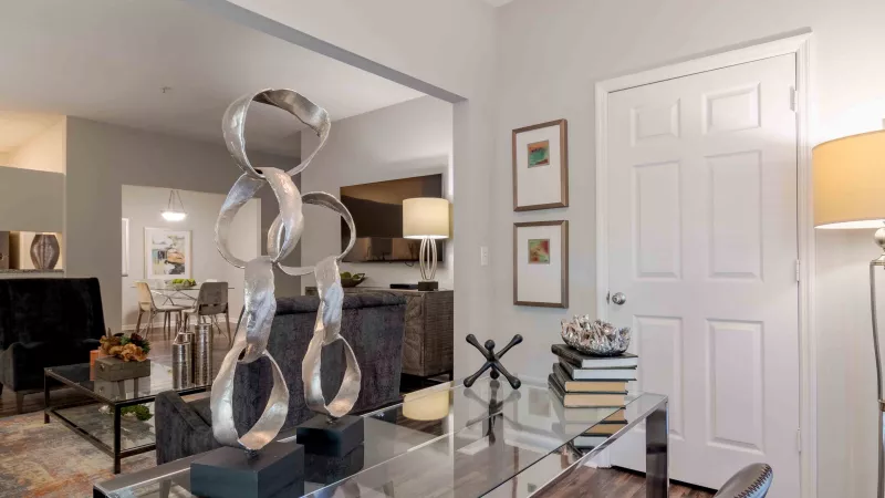 Modern home office with a glass desk, silver abstract sculpture, and books. Two framed artworks hang near a white door.