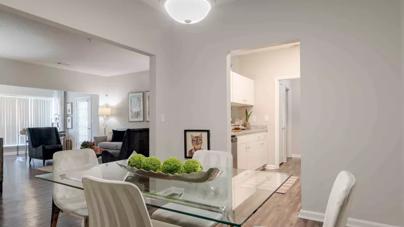 Dining area with glass table and white chairs, leading to a living room and a small kitchen.