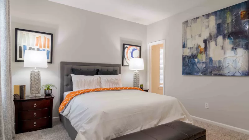 Modern bedroom with a gray tufted king bed, white bedding, orange throw, dark wood nightstands, and abstract art.