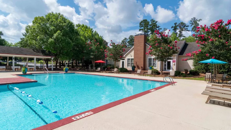 Outdoor swimming pool area with a clubhouse, lounge chairs, and flowering trees under a partly cloudy sky.