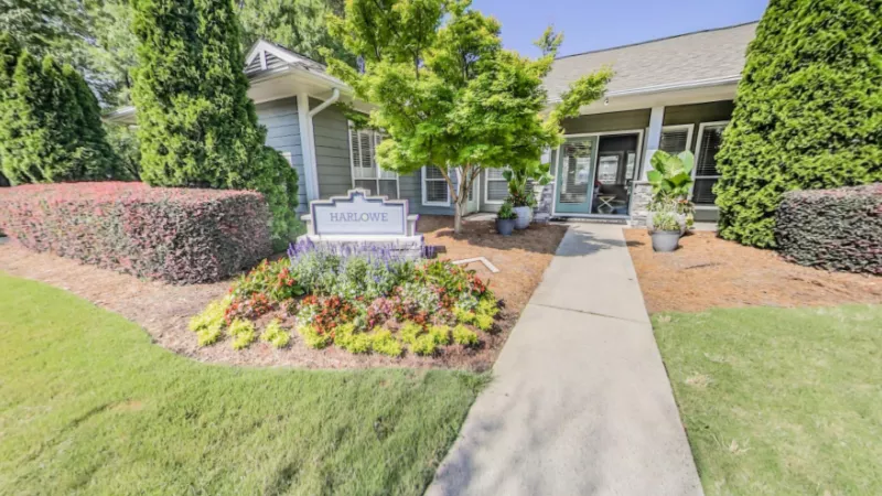 Entrance to The Harlowe Apartments clubhouse featuring a welcoming pathway, colorful flower bed, and neatly trimmed shrubs.