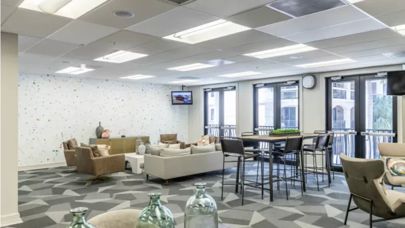 A spacious community lounge with a geometric carpet, modern furniture, and large windows offering ample natural light.