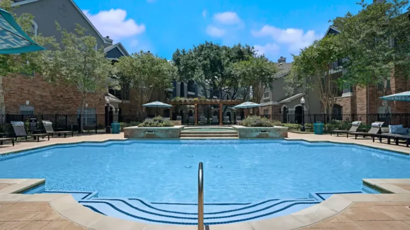 A sparkling blue swimming pool surrounded by lounge chairs and lush greenery at a luxury apartment complex.