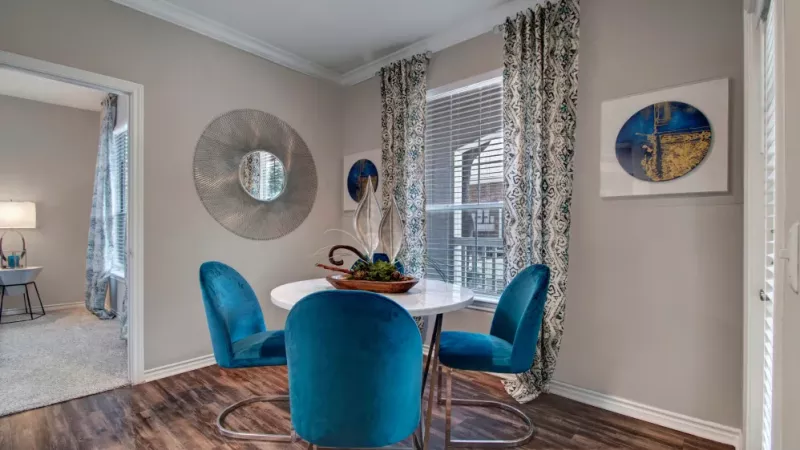 A dining area with vibrant blue chairs, a round white table, and stylish decor, featuring patterned curtains and wall art.