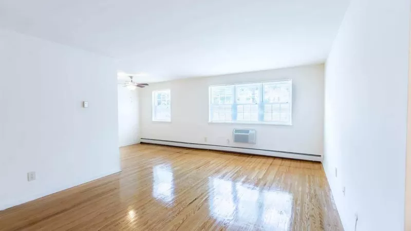 A spacious living room with polished hardwood floors, large windows letting in natural light, and white walls for a fresh and airy feel.