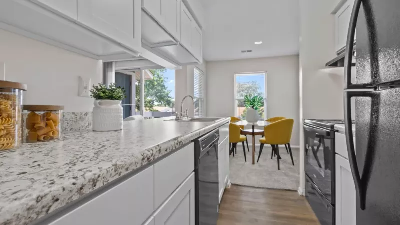 A modern galley kitchen with white cabinetry, black appliances, and a granite countertop leading to a cozy dining area with yellow chairs.