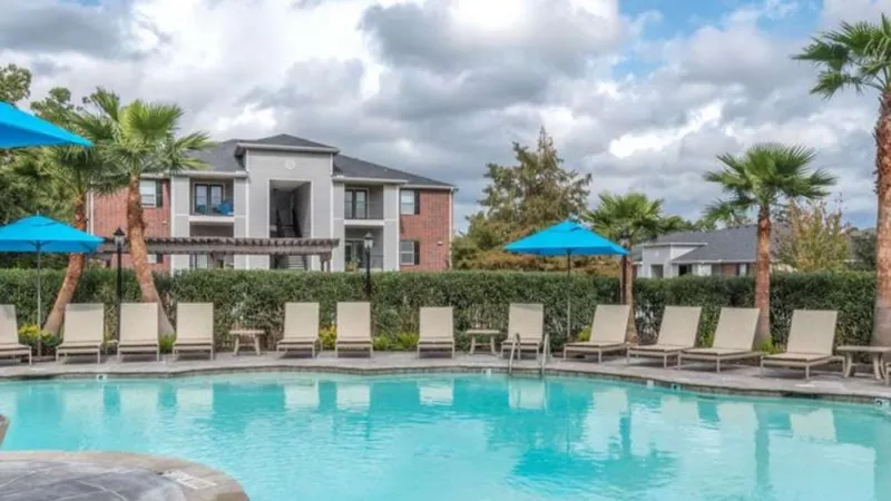 Resort-style swimming pool at The Landings of Conroe Luxury Apartments, surrounded by lush greenery, palm trees, and ample lounge seating under vibrant blue umbrellas.