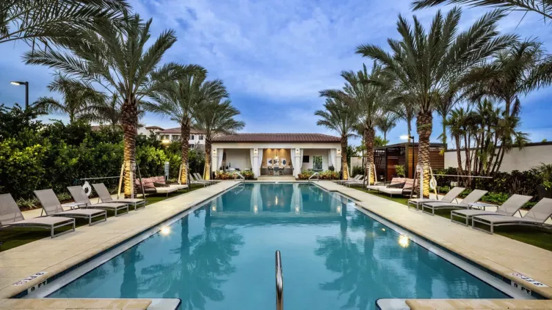 A luxurious swimming pool lined with palm trees and sun loungers, leading to a stylish poolside lounge with cabana seating at Locklyn West Palm Luxury Apartments.