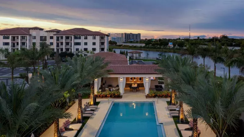 A sunset aerial view of Locklyn West Palm, showcasing the resort-style pool, poolside lounge, and Mediterranean-style apartment buildings with a scenic lake backdrop.