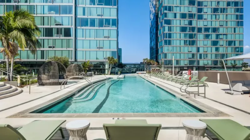 Modern outdoor pool area surrounded by sleek glass apartment buildings with green lounge chairs and palm trees.