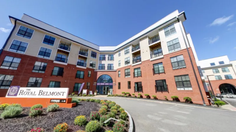 The front entrance of The Royal Belmont Luxury Apartments, showcasing a contemporary red-brick and beige facade with well-maintained landscaping and a prominent community sign.