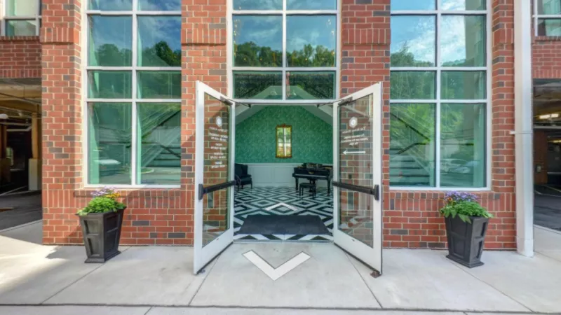 A welcoming entrance featuring a modern brick facade, glass double doors, and elegant planters, leading to a stylish lobby with a grand piano and bold geometric flooring.
