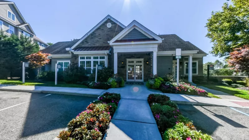 The leasing office at Vista at Town Green Luxury Apartments, featuring a charming stone and siding exterior with a welcoming entrance. The beautifully landscaped walkway is lined with vibrant flowers, creating an inviting atmosphere for residents and visitors.