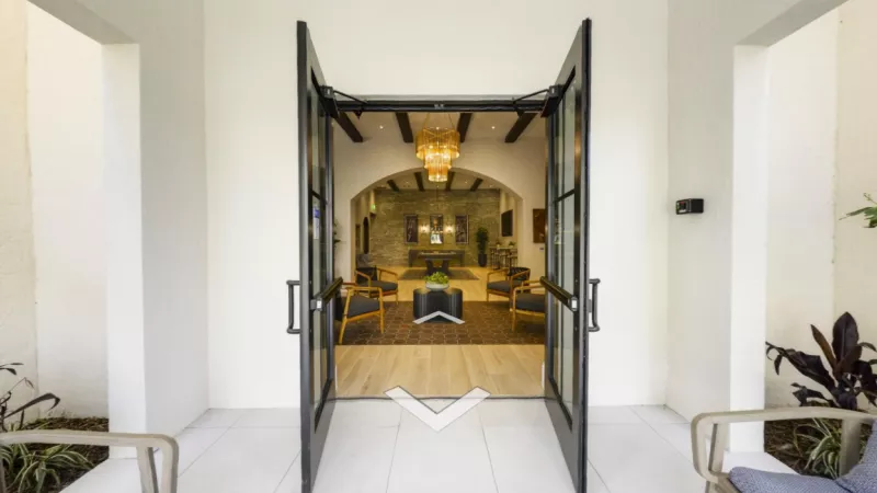 A welcoming entrance to Locklyn West Palm Luxury Apartments featuring stylish black-framed glass doors opening into a beautifully designed lobby with warm lighting, a modern chandelier, and comfortable seating.