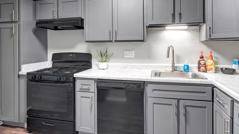 A modern kitchen at Wilson Plaza featuring sleek gray cabinetry with silver handles, a black gas stove with a matching range hood, and a black dishwasher. The kitchen has white marble-style countertops, a stainless steel sink with a high-arc faucet, and under-cabinet lighting. A small potted plant adds a touch of greenery, while soap dispensers and kitchen essentials are neatly arranged on the counter.