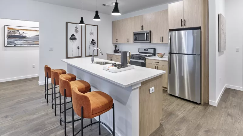 Contemporary kitchen with light wood cabinets, a white quartz countertop island, and three burnt orange bar stools; accented by black pendant lights, stainless steel appliances, and minimalist black-and-white wall art.