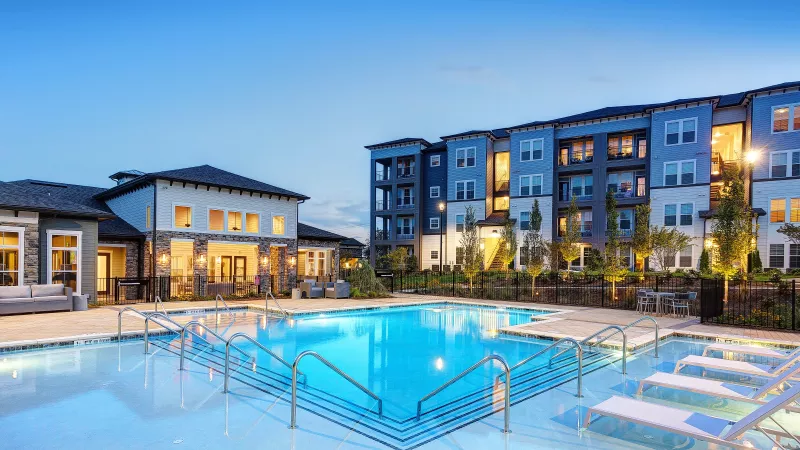 Luxury apartment community with a large resort-style swimming pool, modern lounge chairs, and clubhouse at dusk, with multi-story residential buildings in the background.