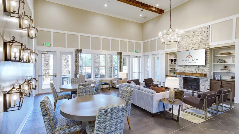 Spacious community clubhouse with high ceilings, exposed wood beams, and elegant lighting fixtures. The lounge includes round dining tables with patterned chairs, multiple seating areas with sofas and armchairs, and a stone fireplace with a mounted TV displaying the Avery at Turkey Creek logo. Built-in shelving, wall sconces, and large windows create a bright and inviting atmosphere.