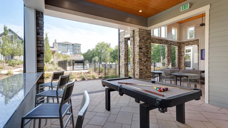 Covered community lounge with a pool table, bar seating, and dining tables, featuring stone accents and views of the landscaped apartment grounds and pool area.