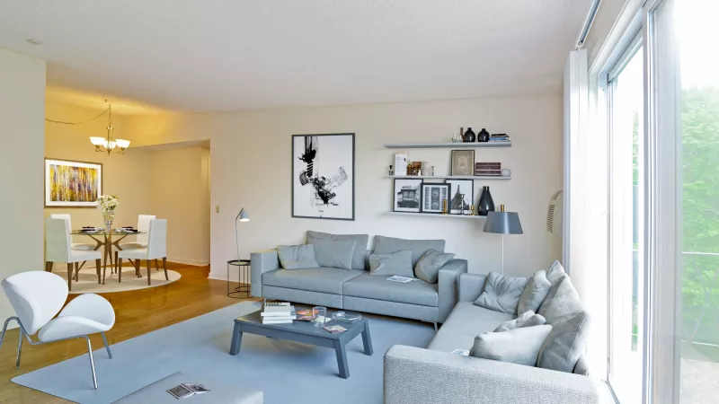 Modern living room with gray sectional sofas, a glass coffee table, and wall art, opening to a dining area with a round table and chandelier, featuring large windows that bring in natural light.