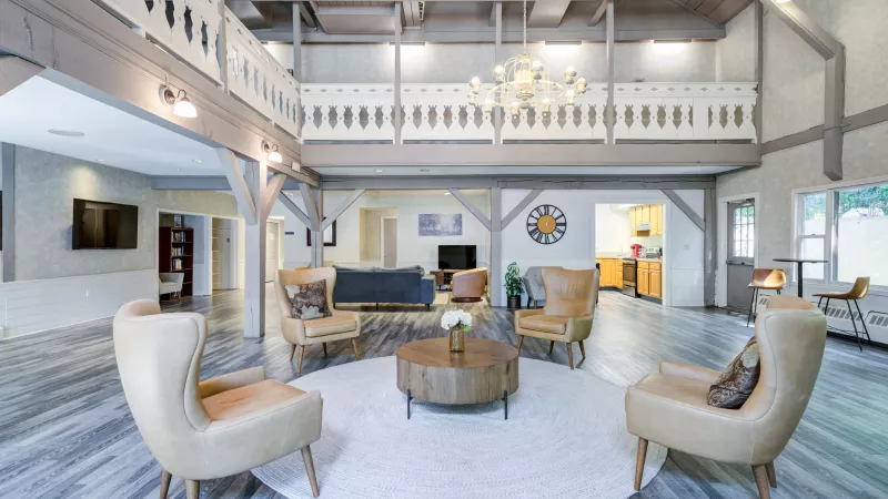 Spacious community clubhouse with vaulted ceilings, exposed beams, and a mezzanine balcony. The room features modern gray wood flooring, a circular white rug, and four tan leather armchairs arranged around a round wooden coffee table. A chandelier hangs above, and the space includes a lounge area with sofas, a mounted TV, a bookshelf, and a kitchen in the background. Large windows bring in natural light, and neutral tones create a warm, inviting atmosphere.