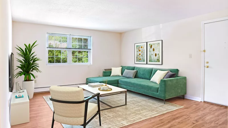 Bright living room with a green sectional sofa, accent chair, coffee table, and wall art, featuring wood-style flooring and a large window that lets in natural light.