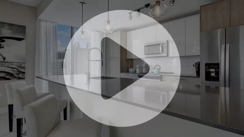 Modern apartment kitchen with stainless steel appliances, pendant lighting, and a large island with bar seating, overlaid with a play button icon indicating a virtual tour preview.