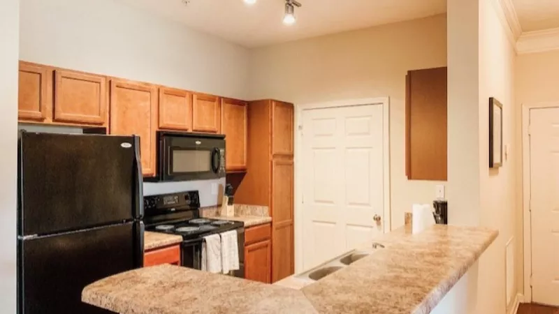 Modern apartment kitchen with wood cabinets, black appliances, and granite-style countertops under warm lighting.