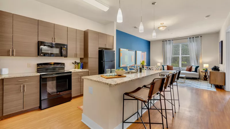 Modern open-concept kitchen and living area featuring wood-style flooring, granite countertops, and contemporary brown cabinetry with stainless steel handles. The kitchen includes black appliances, pendant lighting over a breakfast bar with two leather barstools, and a view into a cozy living space with a blue accent wall, white sofa, artwork, and large window with sheer curtains.